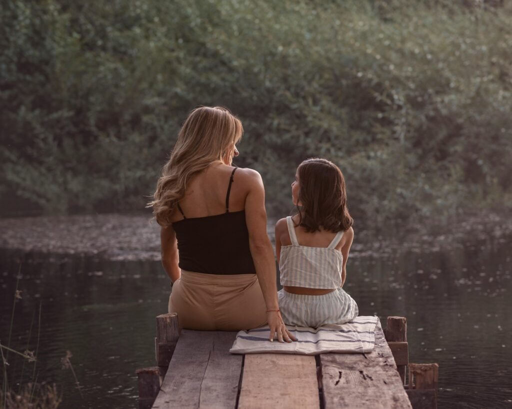 Mother and daughter sitting together on a wooden dock by the water at sunset, sharing a quiet moment of connection and conversation