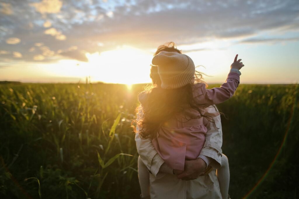 Mother holding her daughter in a sunlit field at sunset, looking out together and sharing a quiet, comforting moment