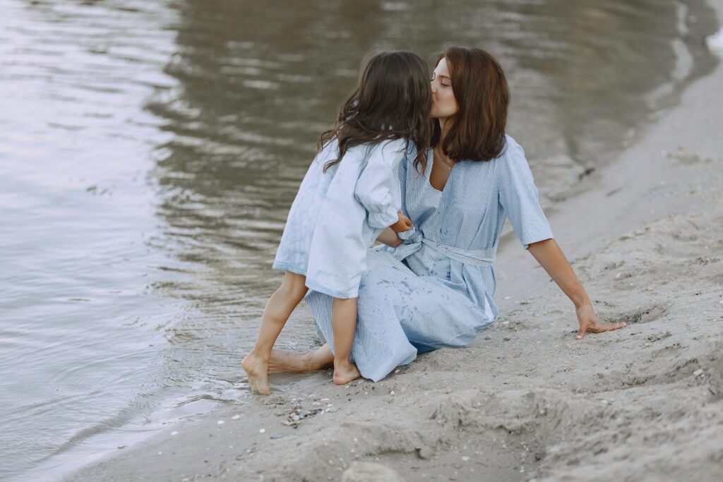 Mother sitting on the sand by the water, gently kissing her daughter as they stand close together at the shoreline.