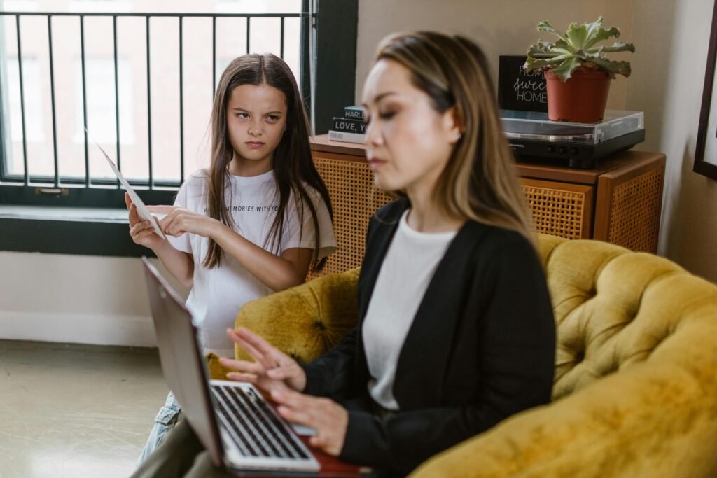 Child standing by the couch looking frustrated while her parent focuses on a laptop, showing tension and disconnection.